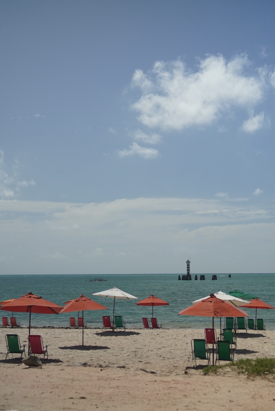 Guarda-sóis coloridos na areia, com o mar e um farol ao fundo